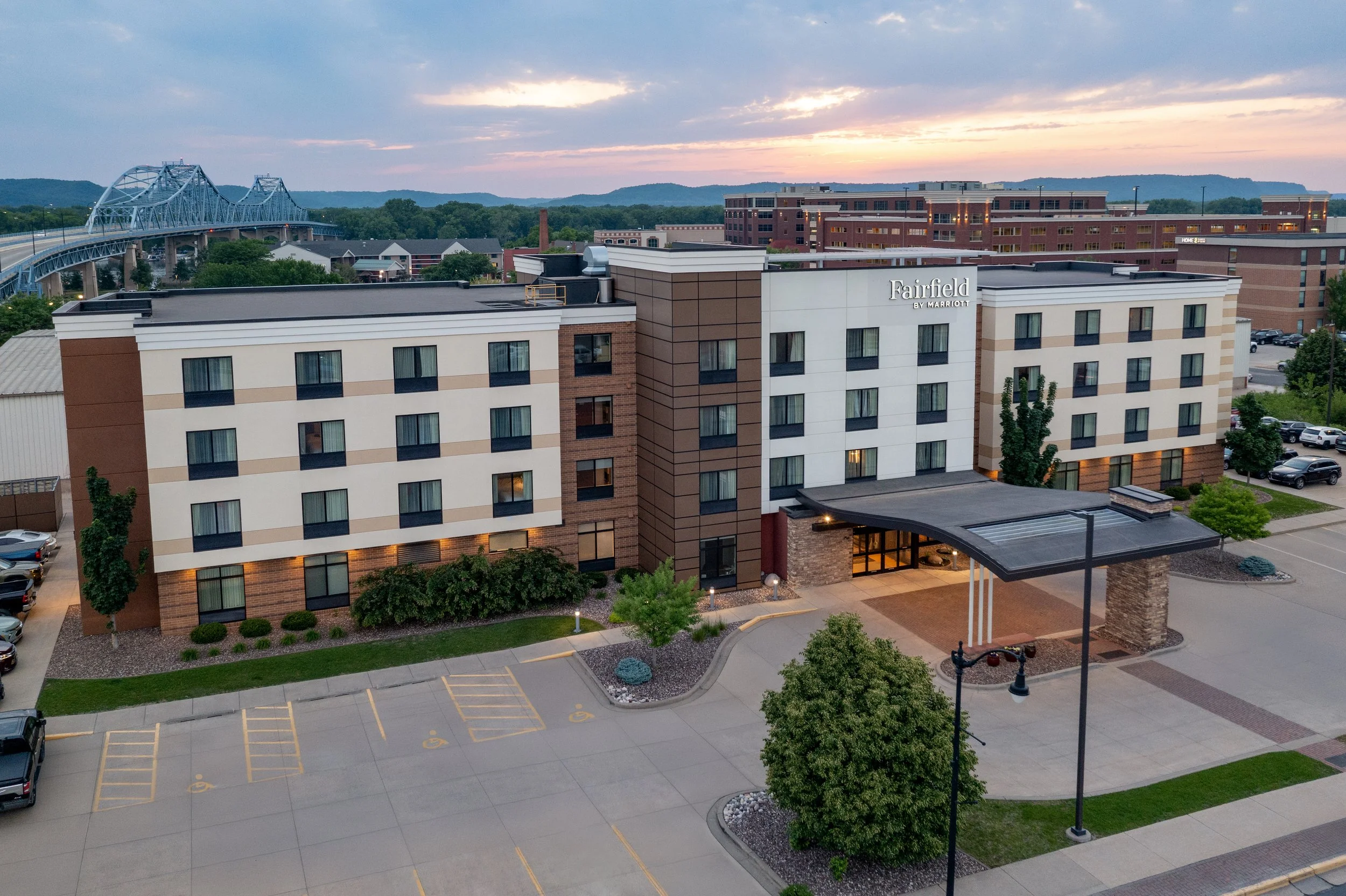 An aerial view of the Fairfield by Marriott hotel with parking lot in foreground, trees, and a bridge over a river in the background during sunset.