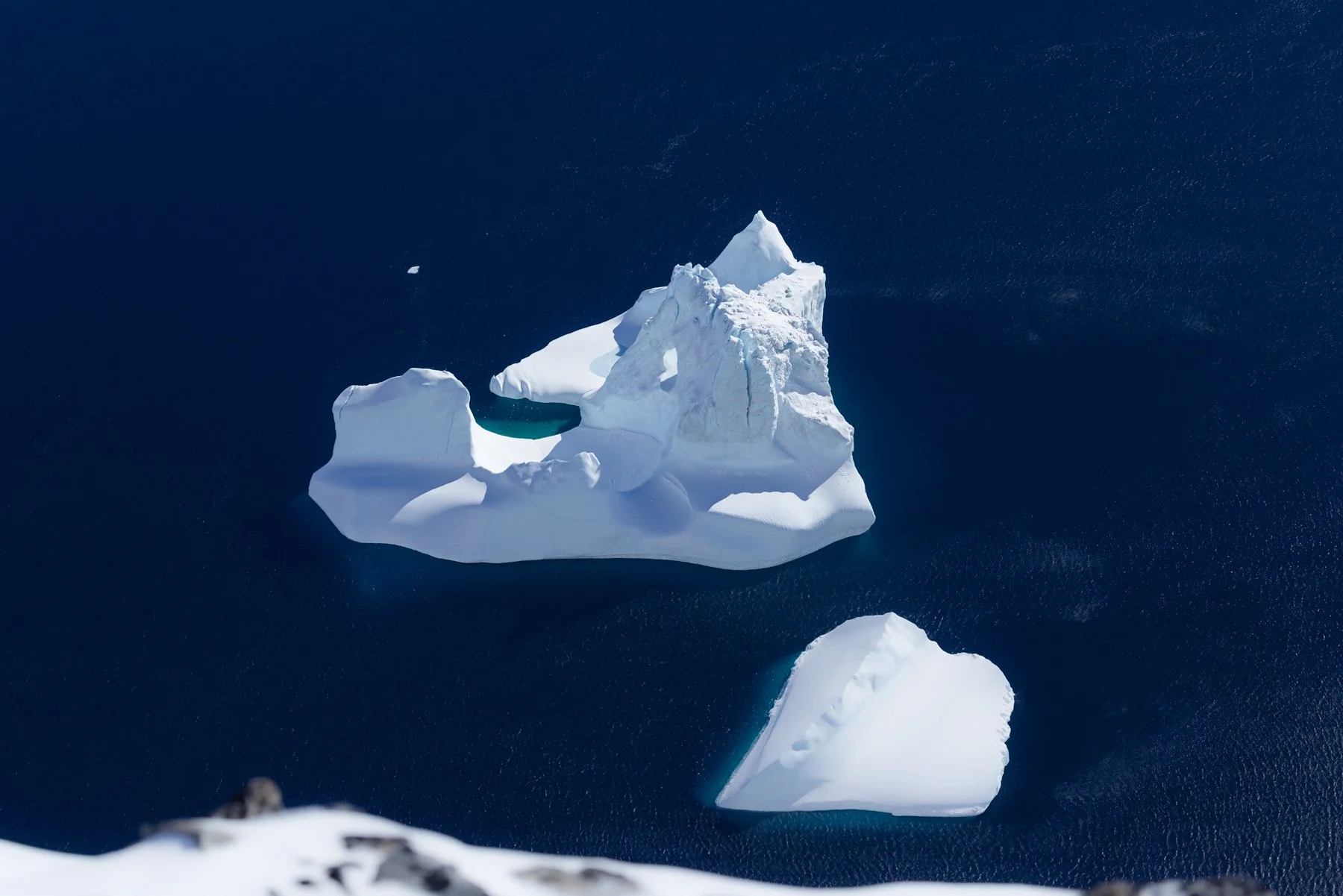 Eine große Eisscholle im tiefblauen Wasser, mit einer kleinen Eiskuppel auf der Oberfläche, aufgenommen aus der Vogelperspektive.