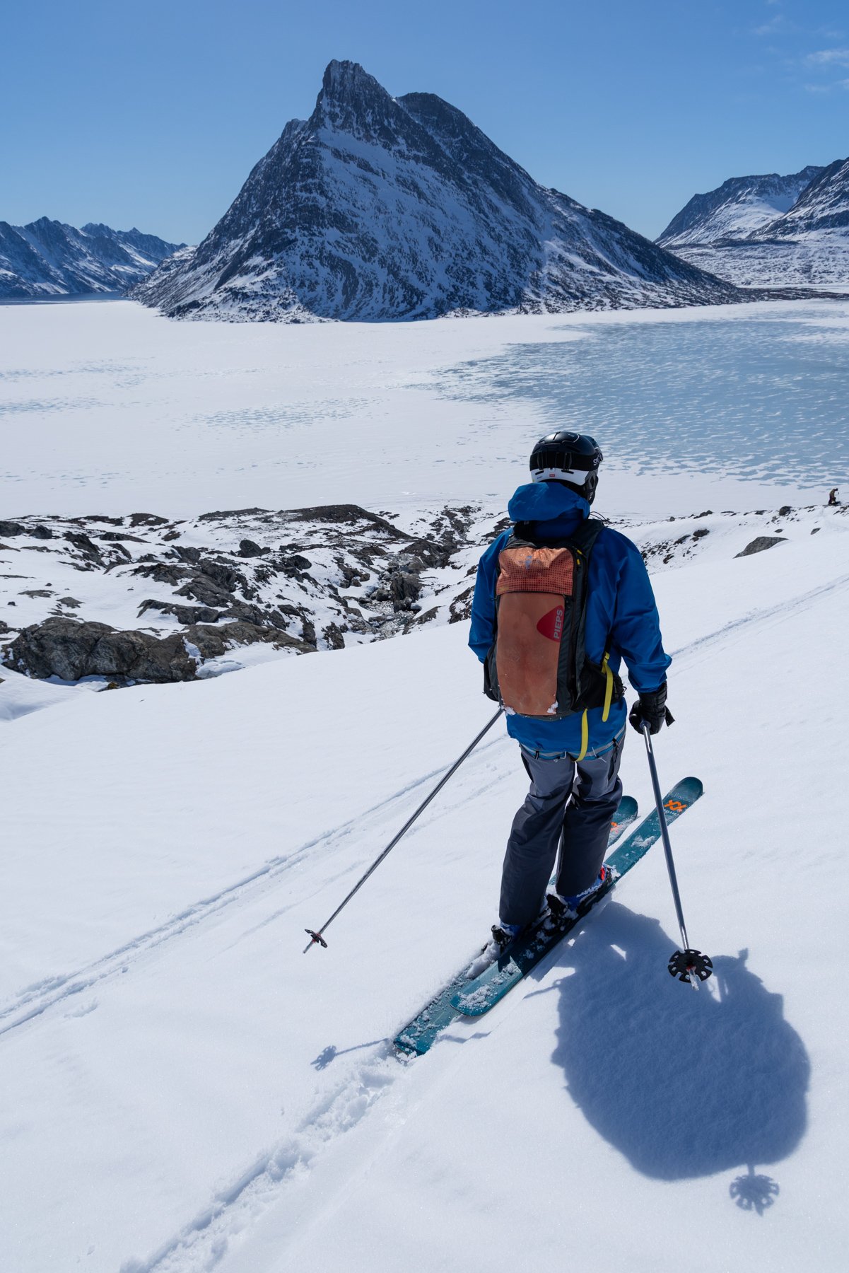 Ein Skifahrer in blauem Kleid und mit Rucksack steht in schneebedeckter Landschaft mit Bergen im Hintergrund.