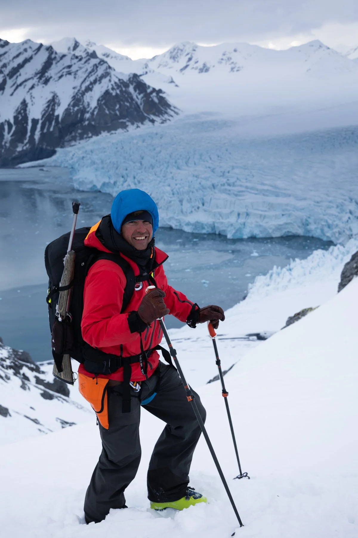 Ein Mann in roter Jacke und blauer Hose mit Wanderrucksack und Wanderstöcken im Schnee, trägt Sonnenbrille und Mütze.
