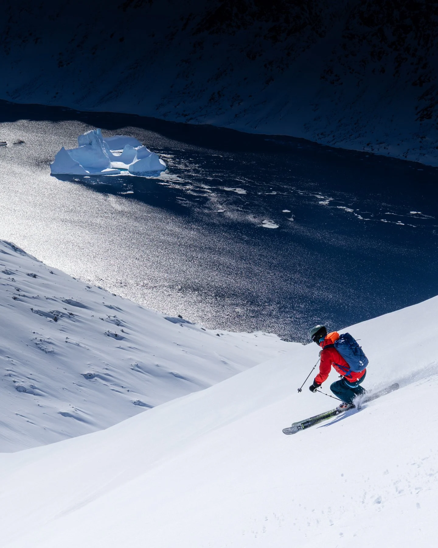 Ein Skifahrer in roter Jacke und blauer Hose fährt eine schneebedeckte Abfahrt hinunter in einer verschneiten Berglandschaft, im Hintergrund flache Eisberge und dunkle Berge unter einem bewölkten Himmel.