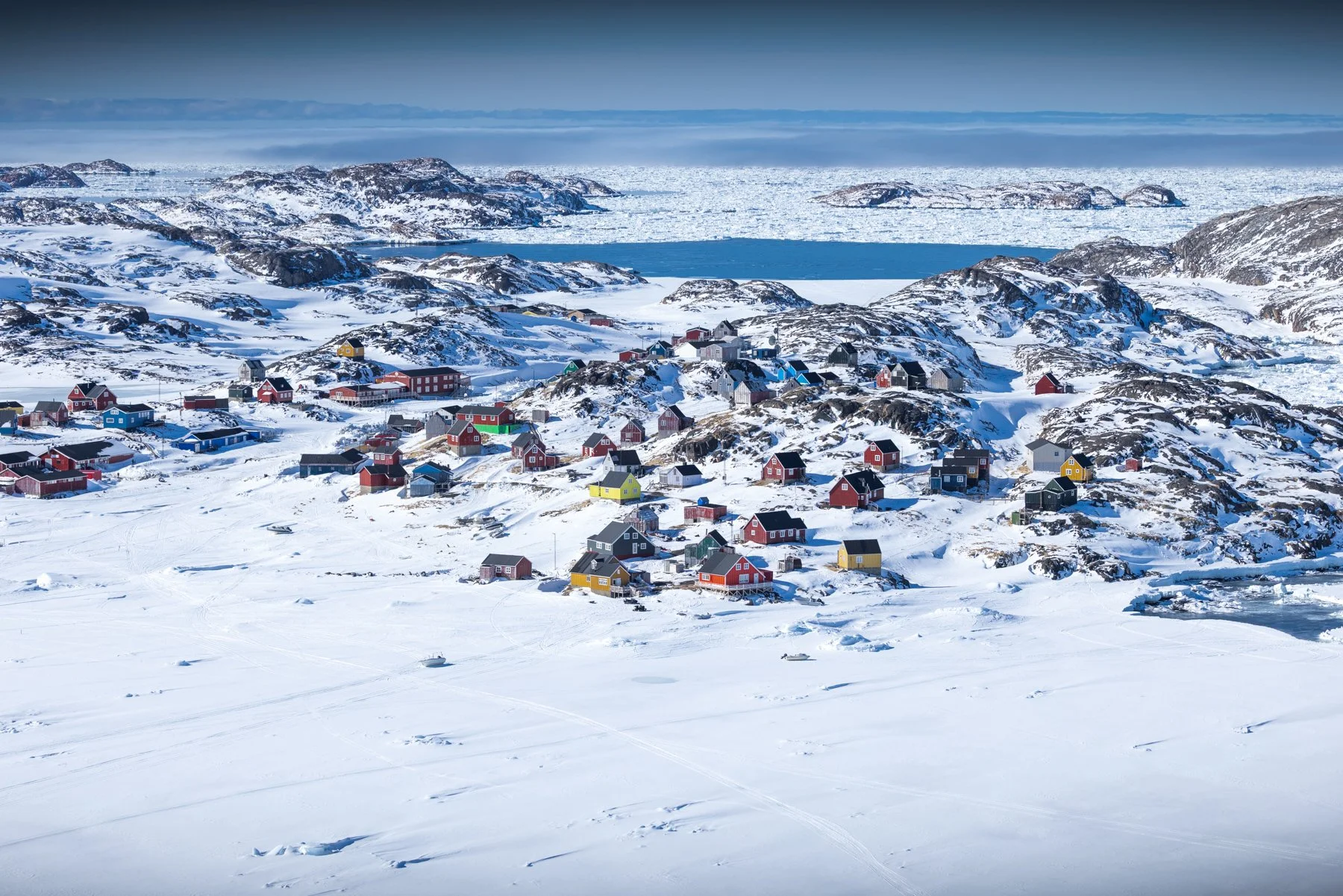 Bunte Häuser in einer verschneiten arktischen Landschaft mit eisbedecktem Wasser im Hintergrund.