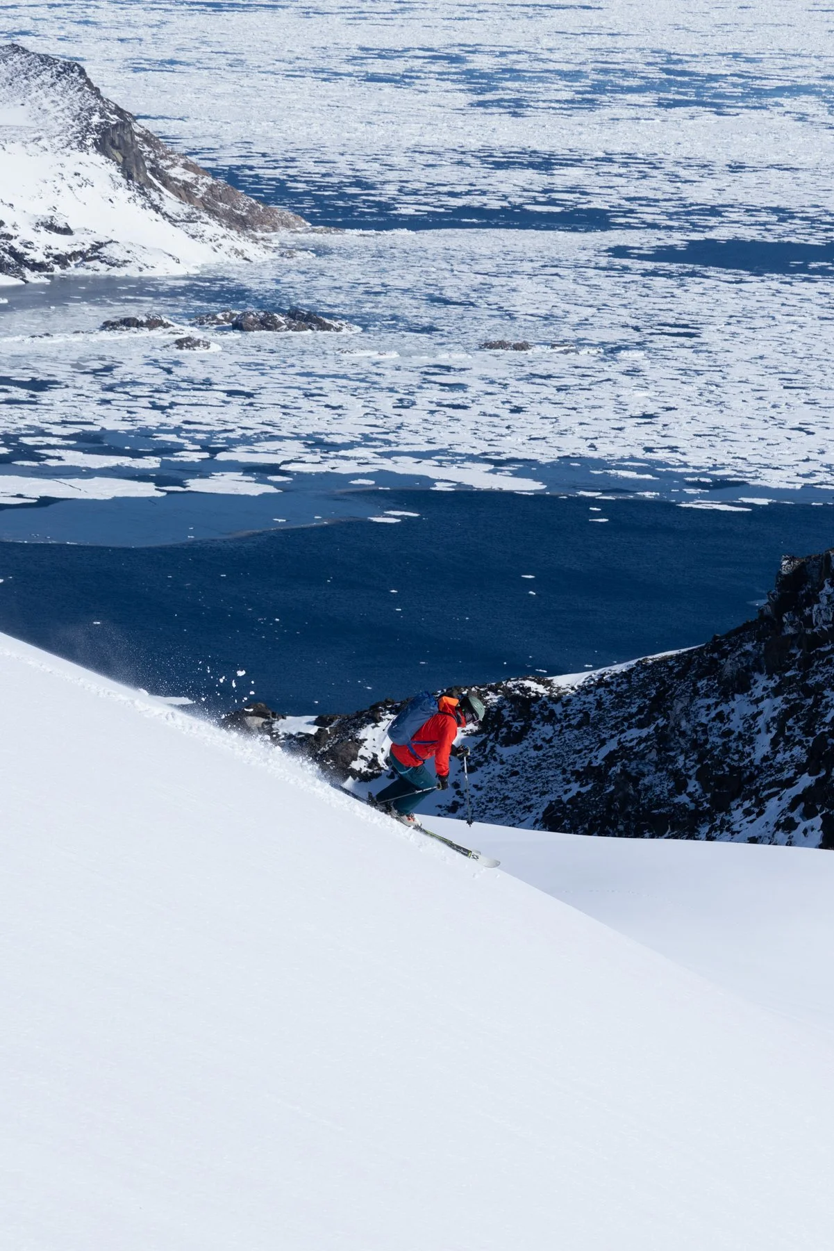 Ein Skifahrer in roter Jacke fährt an einer verschneiten Bergseite ab, im Hintergrund ein gefrorener See mit Eisschollen, umgeben von schneebedeckten Bergen.