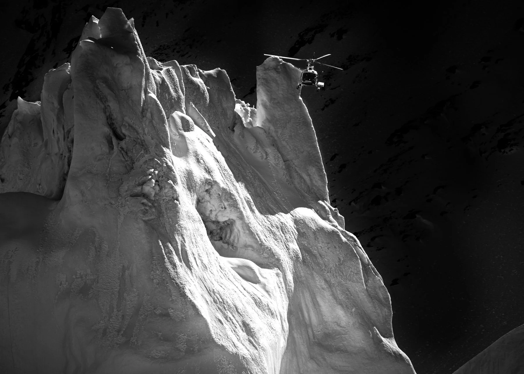 Ein Helikopter fliegt über eine große, schneebedeckte Gletscherklippe in einer alpinen Umgebung bei Dunkelheit oder im Schatten.