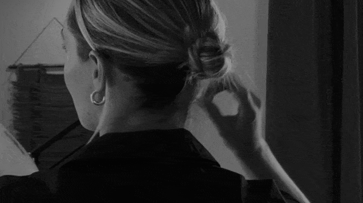 Back of a woman with her hair in a bun, holding her right hand near her ear, in a black-and-white photo.