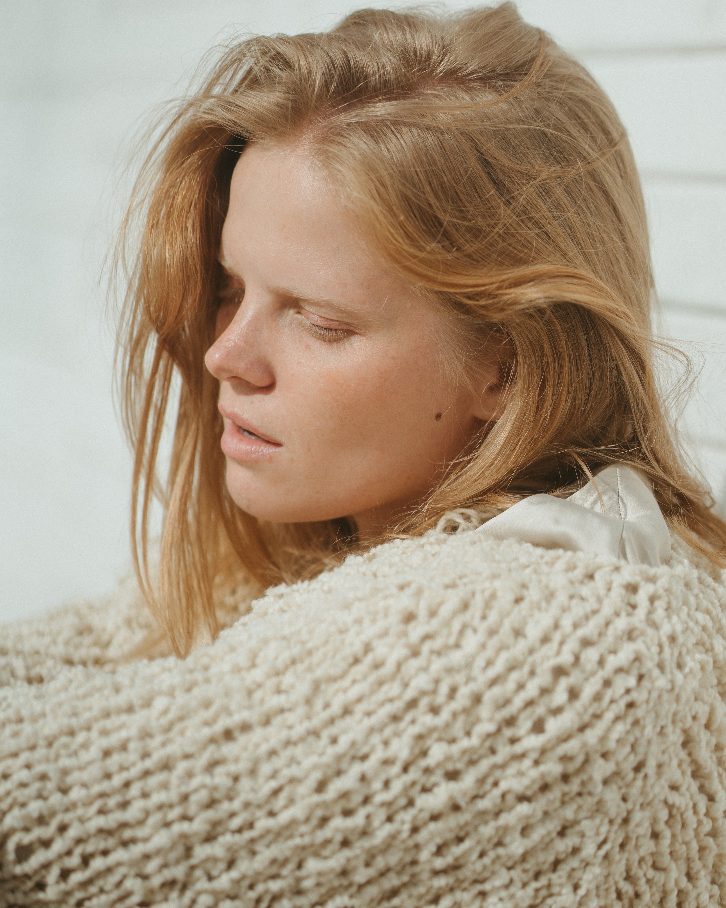Close-up of a red-haired woman with her eyes closed, wrapped in a beige knitted blanket, against a white background.