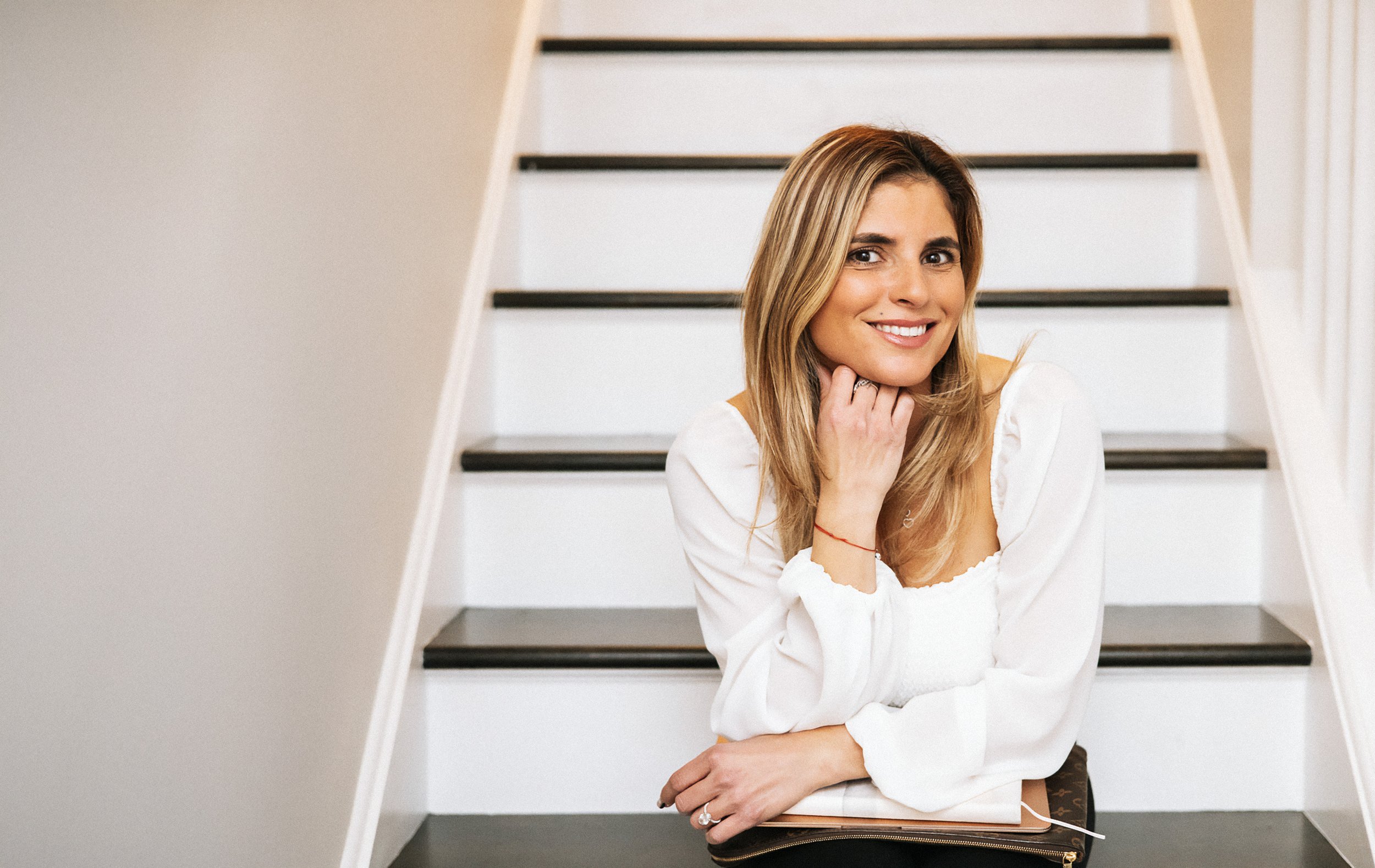 A woman with blonde hair and a white blouse sitting on stairs, smiling at the camera.