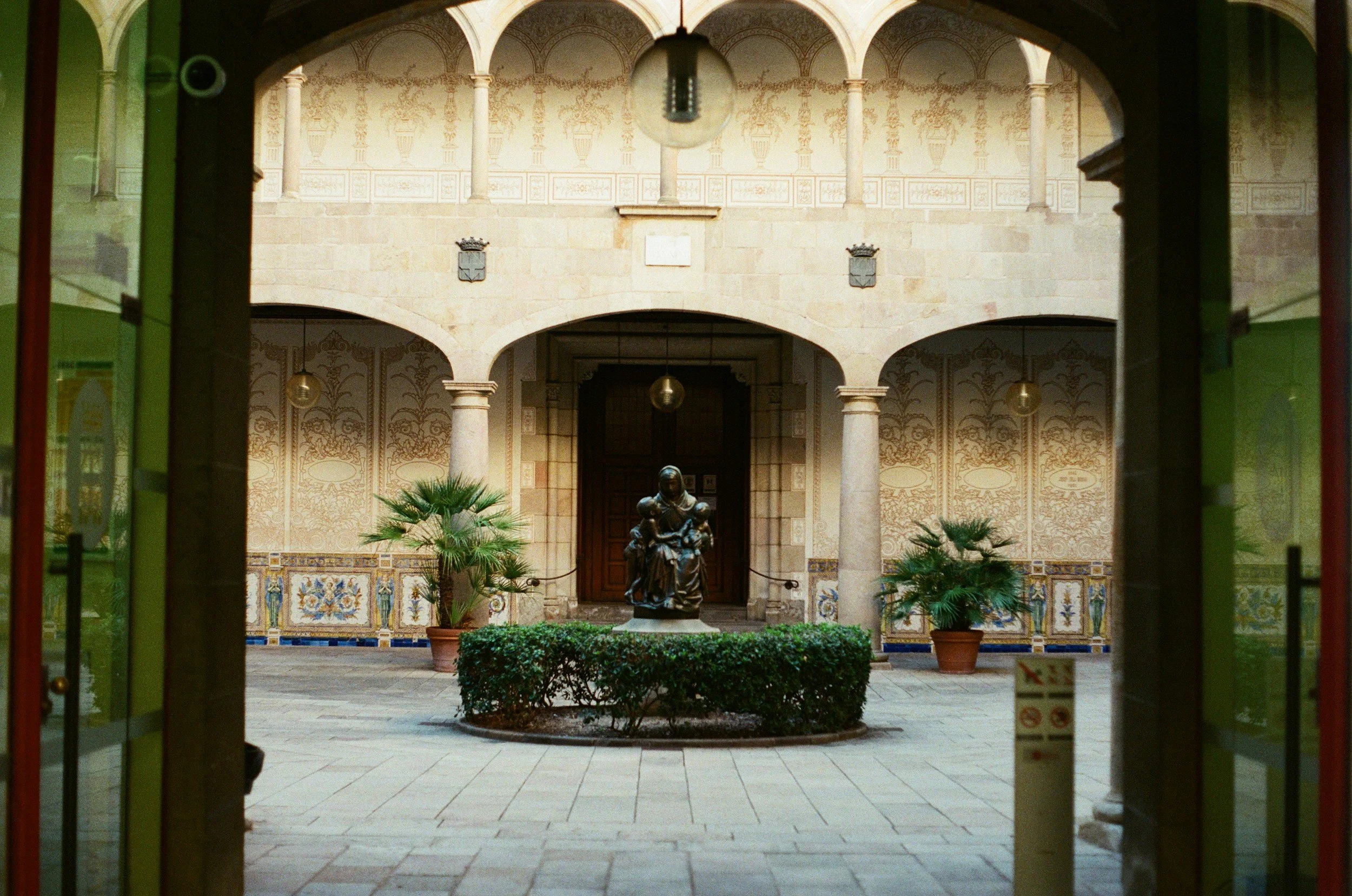 View of an elegant courtyard through a doorway with green frames, featuring a statue of a woman holding a child, potted plants, and ornate tiled walls with crowned shields.