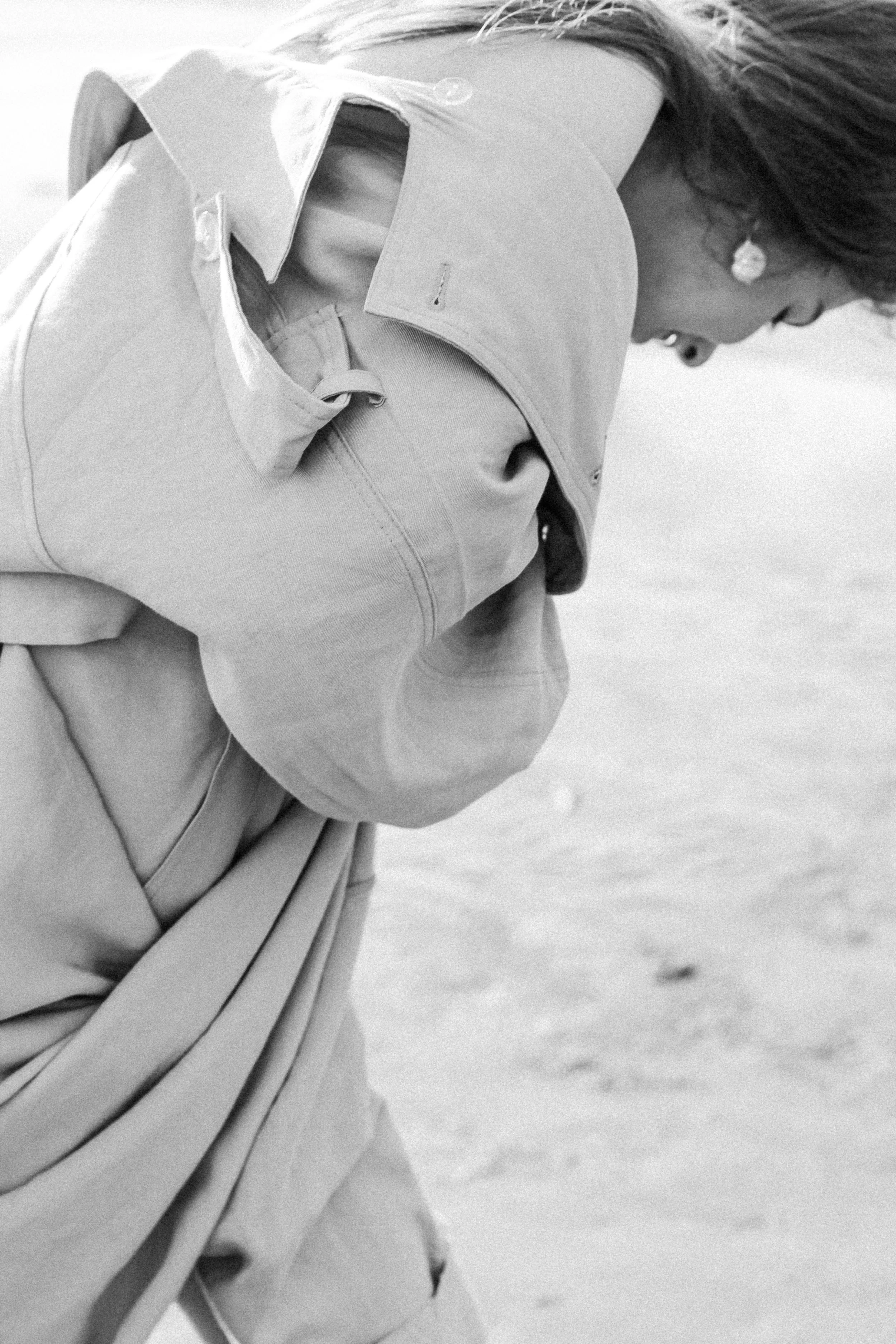 A woman in a light-colored jacket and pants, wearing earrings, bent over near a body of water, captured in a black and white photograph.