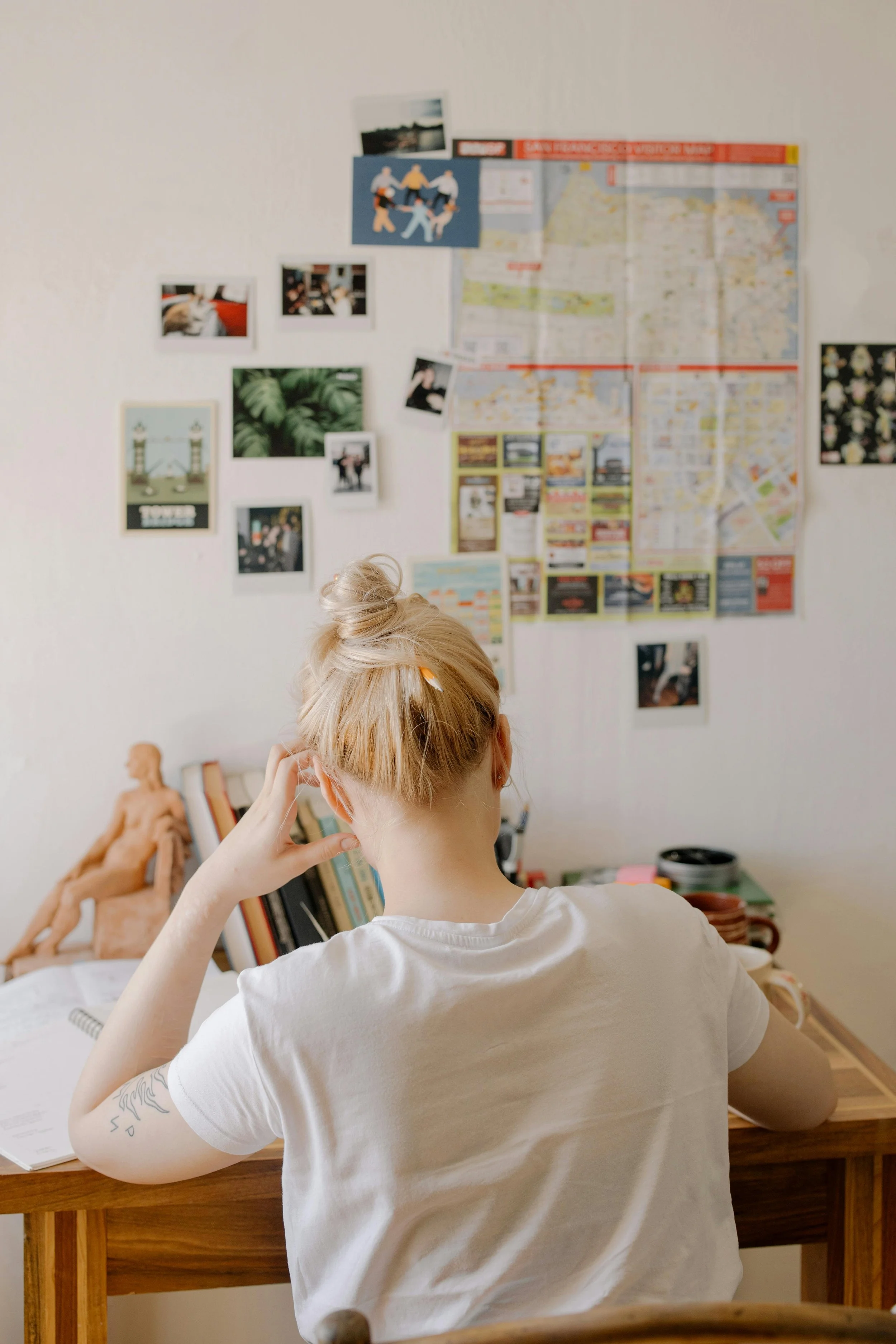 A person with blonde hair in a bun, wearing a white t-shirt, sitting at a wooden desk, looking at a wall decorated with various photos, maps, and notes.