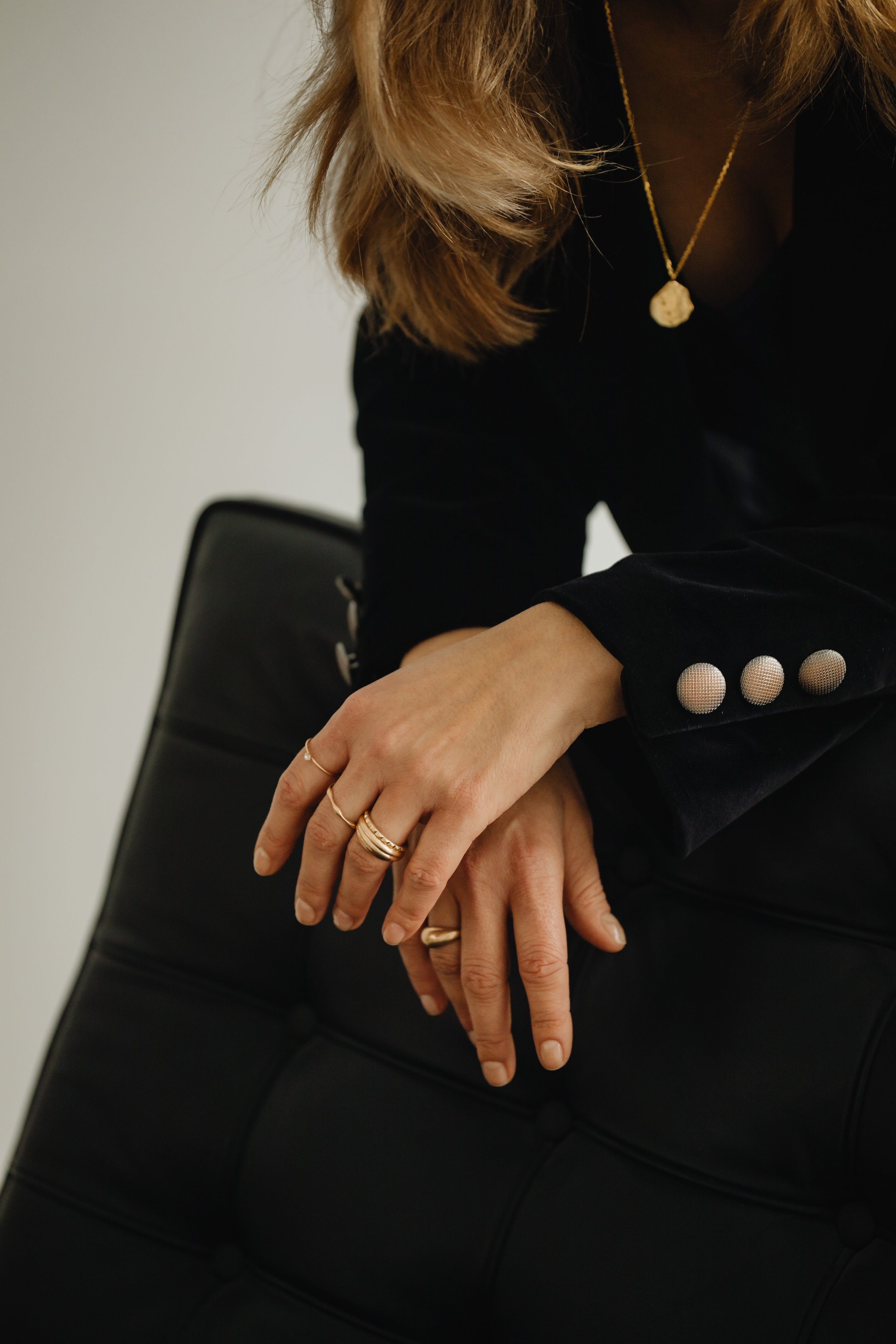Close-up of a woman with blonde hair wearing rings and a black blazer, sitting on a black chair.