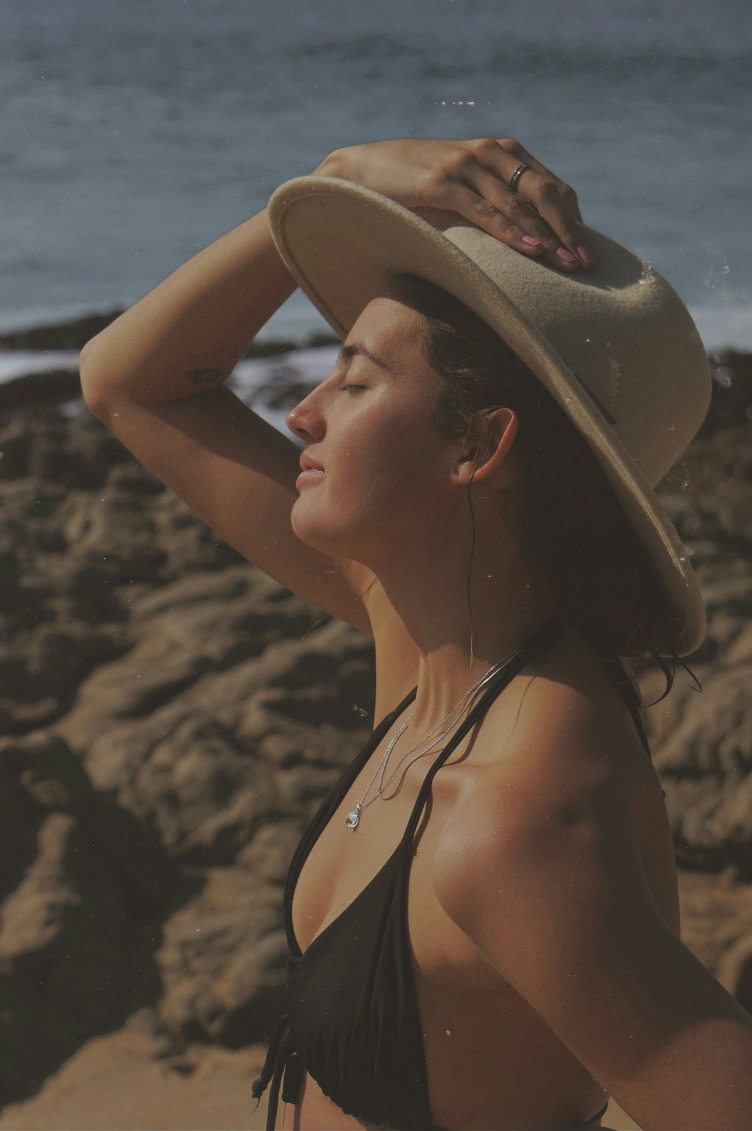 A woman at the beach wearing a black swimsuit and a wide-brimmed hat, with her eyes closed and sunlit face, holding her hat with one hand, and wearing earphones and a delicate necklace.