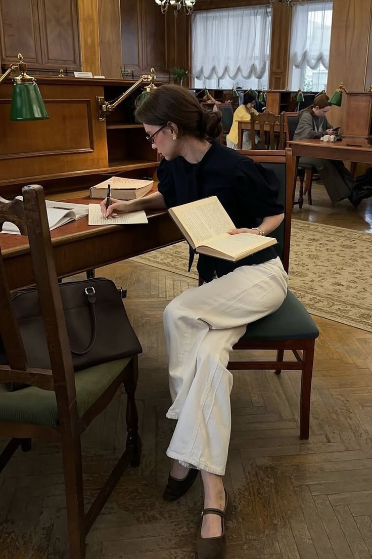 A woman with dark hair tied in a bun, wearing glasses, a black top, white pants, and black heels, is sitting at a wooden desk in a library. She is writing in a notebook, holding a book in her left hand, with several open books on the desk. Two other people are visible in the background, sitting at separate desks, in a room with wood-paneled walls, green desk lamps, and large windows with curtains.