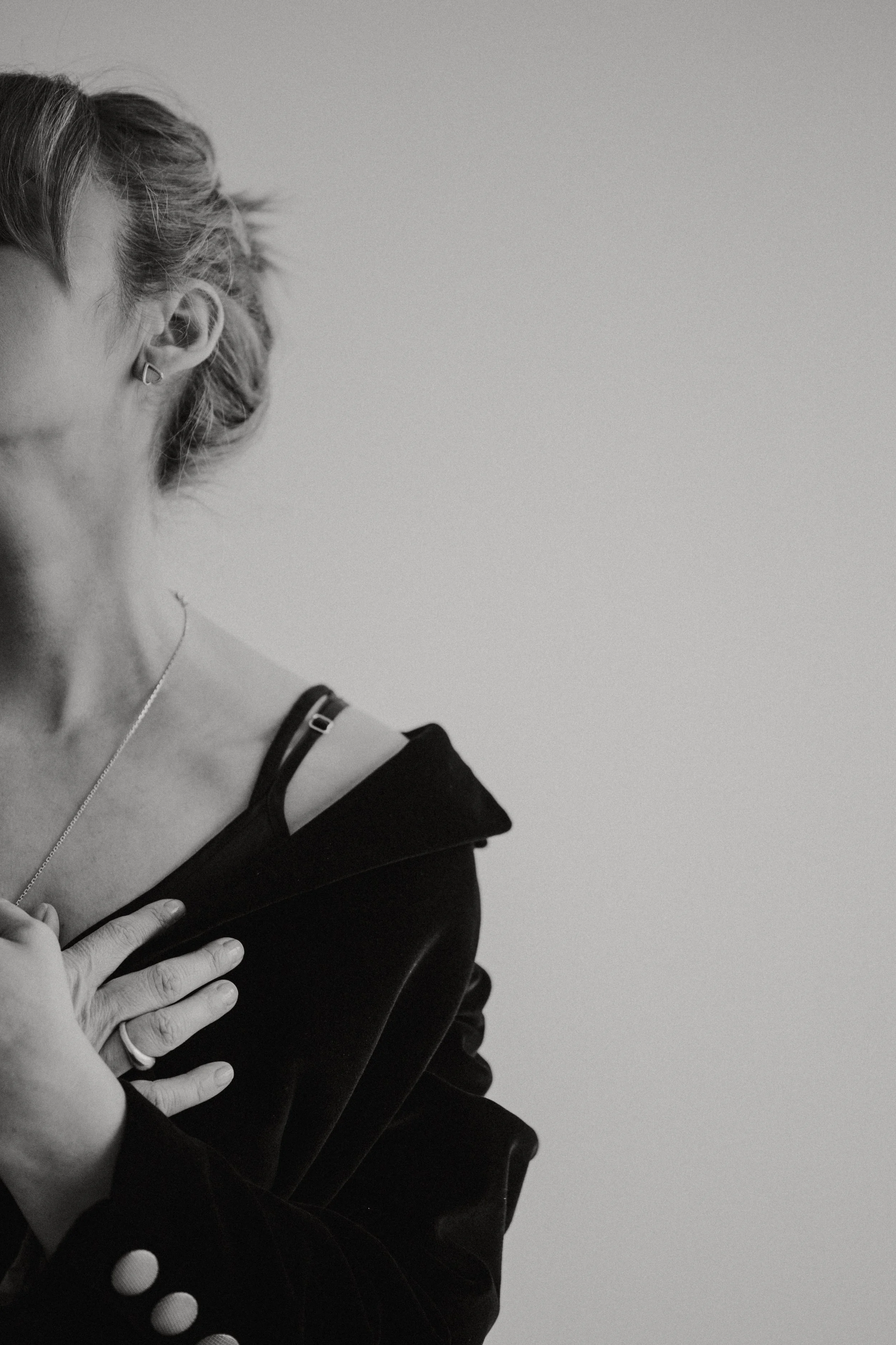 Black and white photograph of a woman with short hair, wearing a ring, a necklace, a black jacket, and a black strap top, with her hand on her chest.