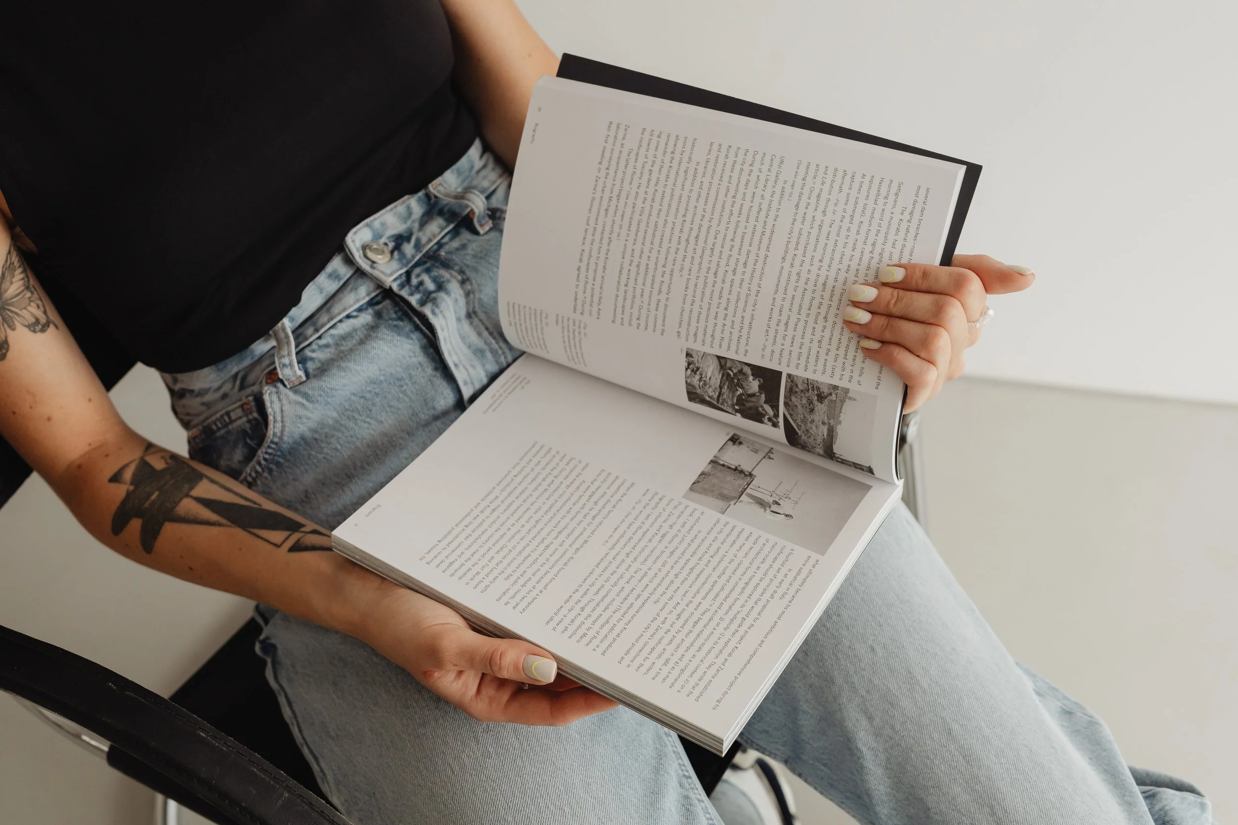 Person sitting on a chair, reading a magazine, wearing light blue jeans and a black shirt, showing tattoos on their arm, with a neutral background.