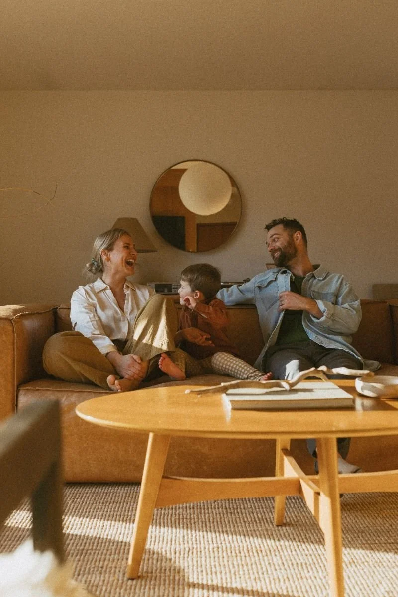 Family of three sitting on a couch, laughing and playing together in a cozy living room.