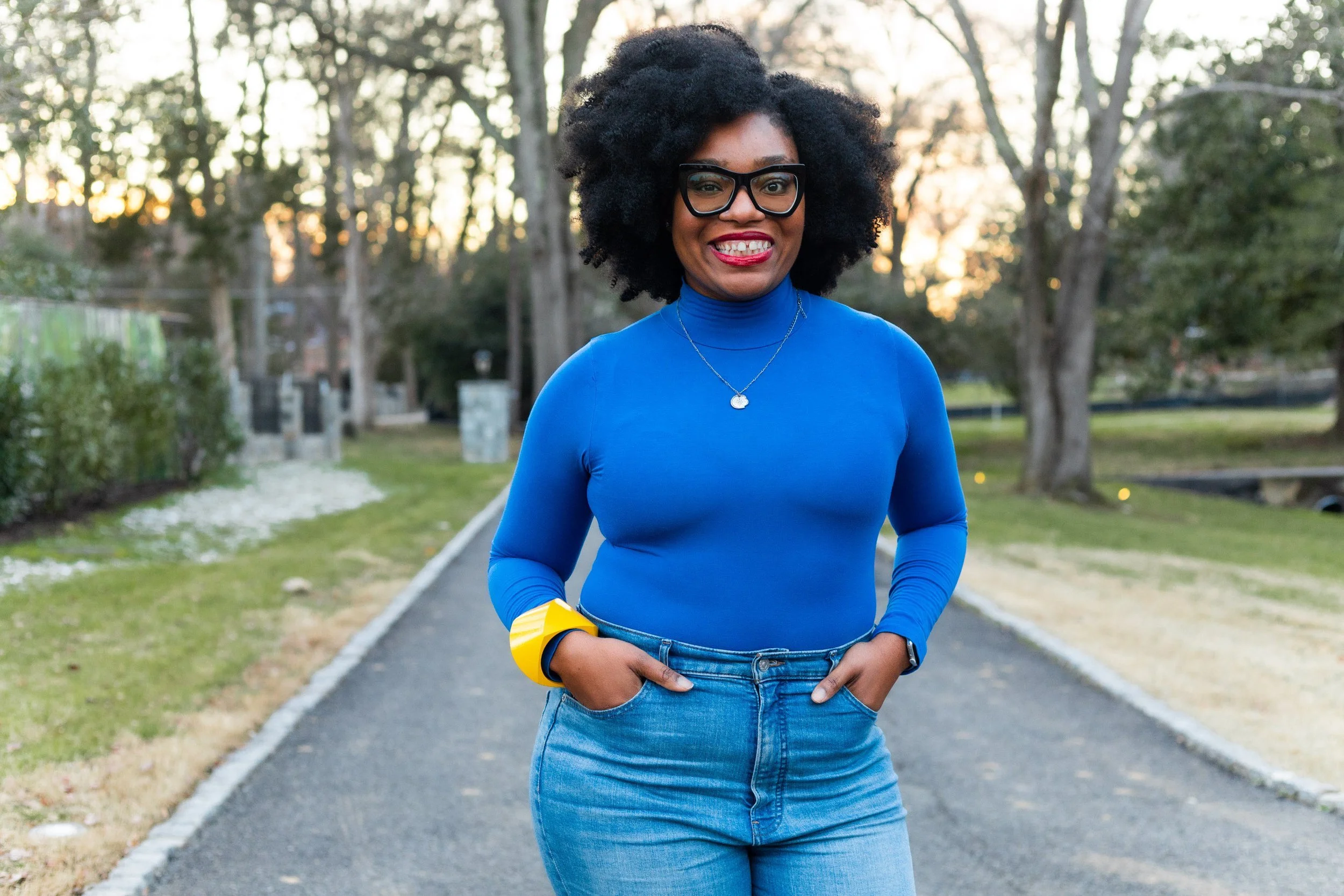 Woman with curly black hair wearing glasses, a blue long-sleeve top, and jeans, smiling while standing outdoors on a park pathway during sunset.