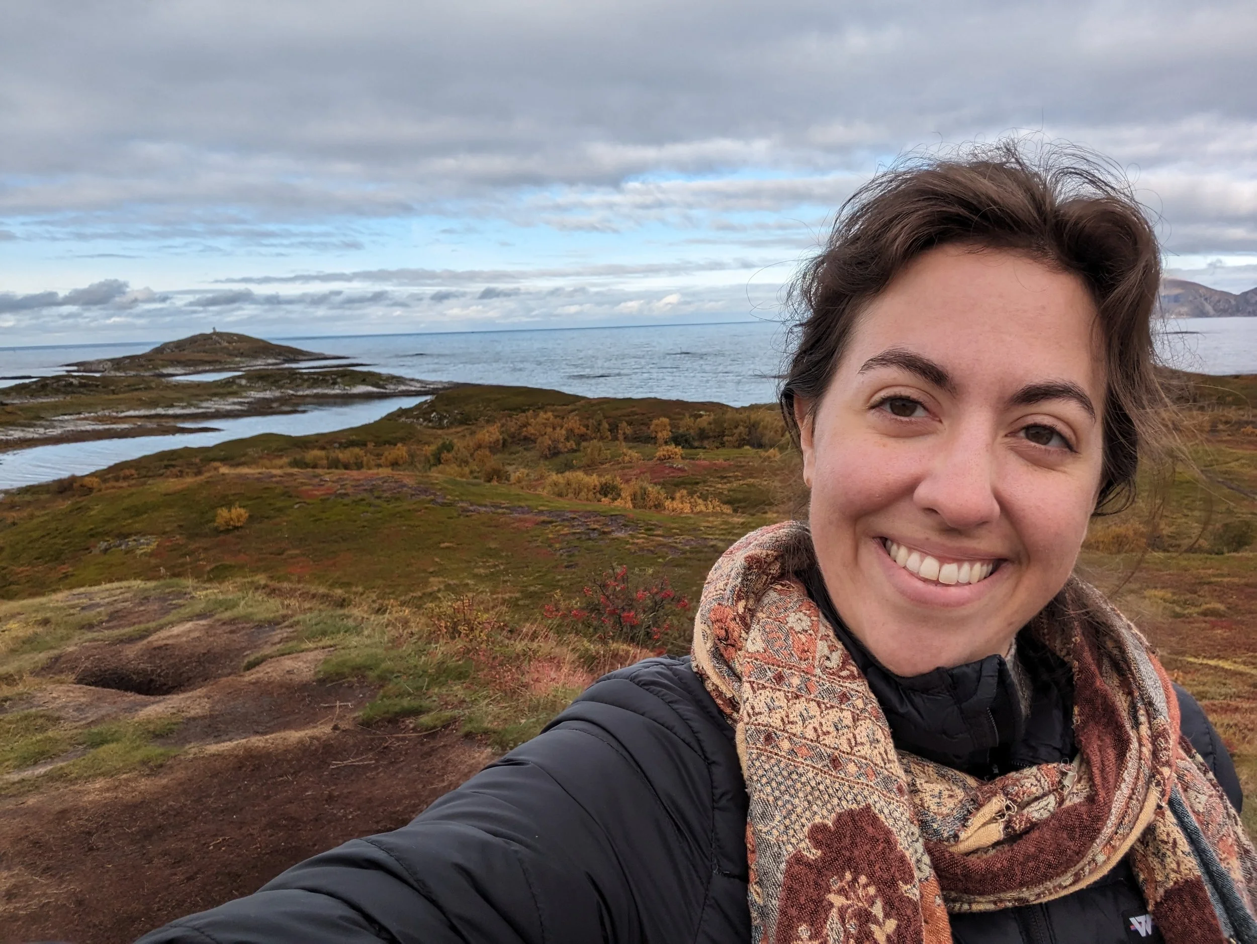 A photo of Abby standing outside in the fall time at the seashore in Norway wearing a black jacket and a multi-colored scarf.