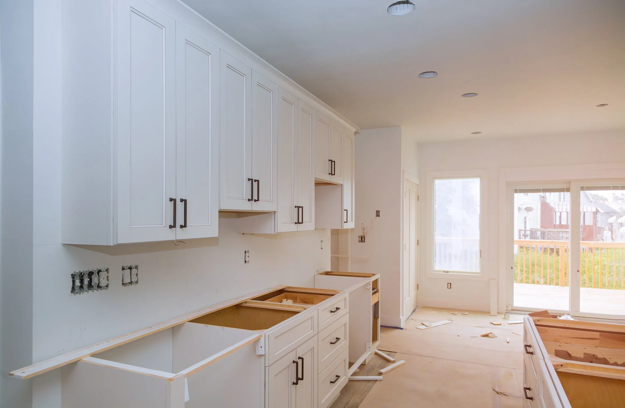 Kitchen under construction with white cabinets and uninstalled countertops, construction debris on the floor, and large windows with a view of an outdoor balcony.