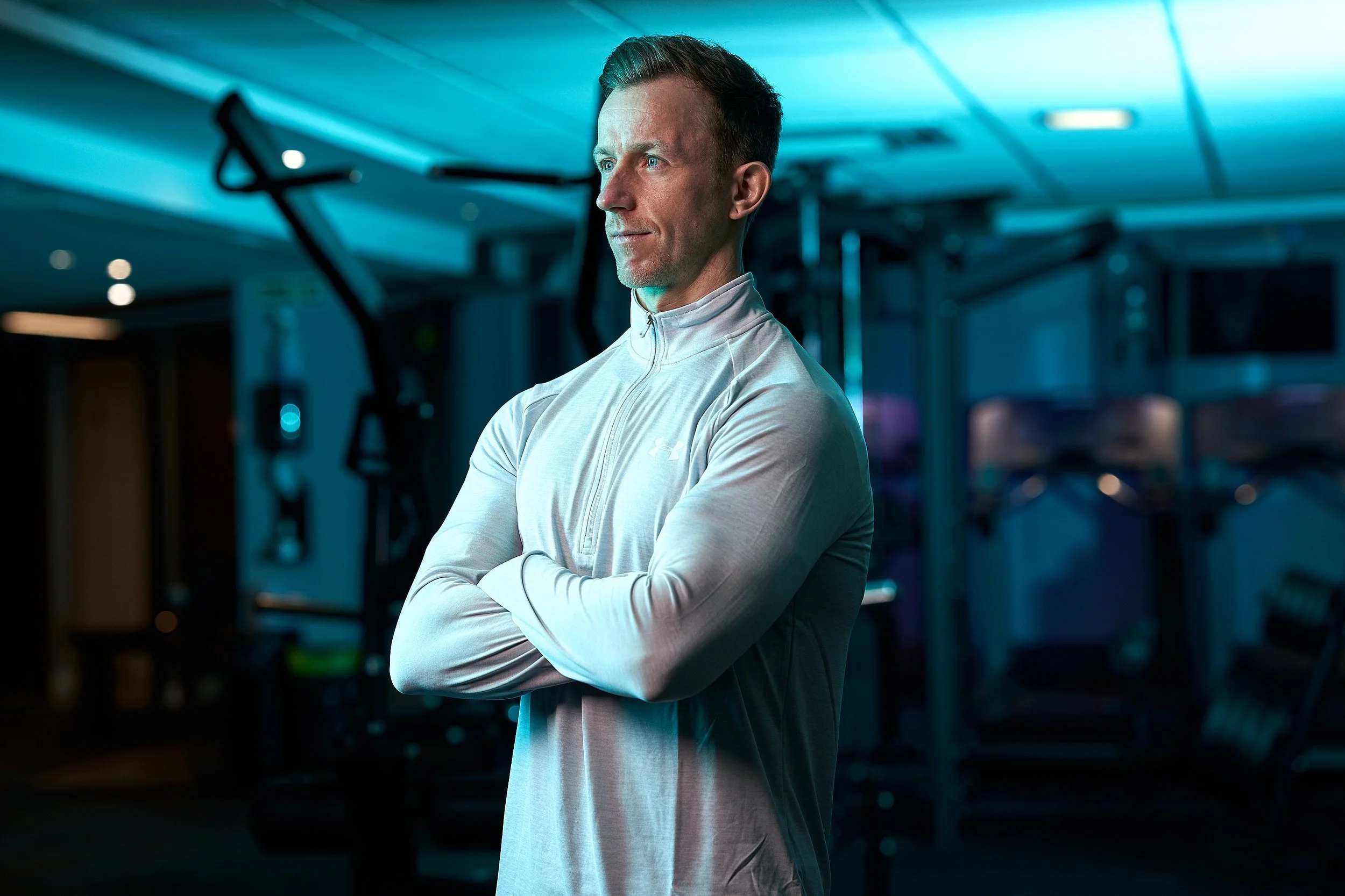 A personal trainer standing with arms crossed in a gym, with weightlifting equipment in the background.