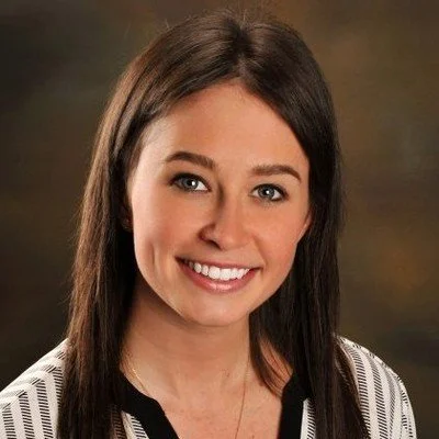 A smiling woman with straight brown hair, wearing a black and white striped top, against a blurred brown background.