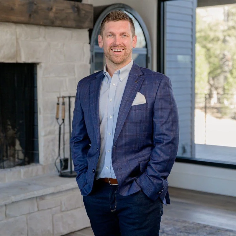 A man with short brown hair and a beard, wearing a blue checked blazer, light blue dress shirt, and dark blue pants, standing indoors in front of a fireplace with tools and large window with a view of trees.