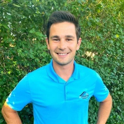 A young man smiling outdoors in front of green foliage, wearing a bright blue collared shirt with a logo on the chest.