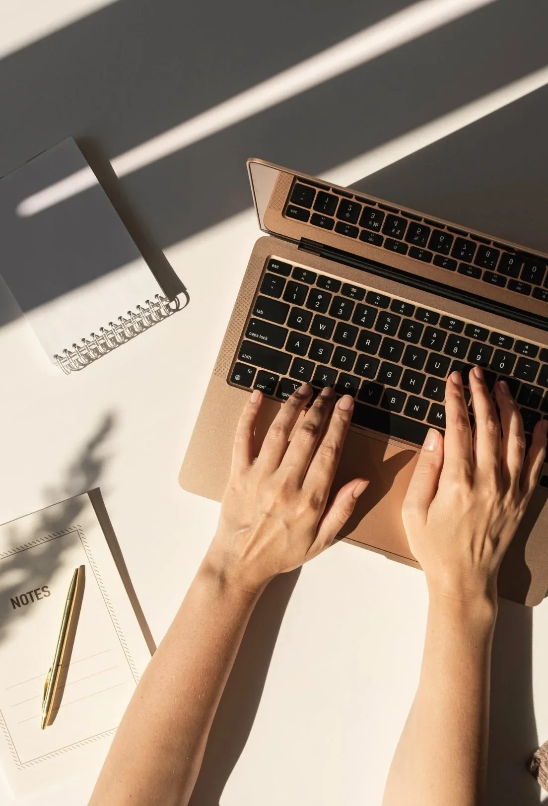 Overhead view of person typing on a laptop with a notepad, pen, and ruler on a white desk in sunlight.