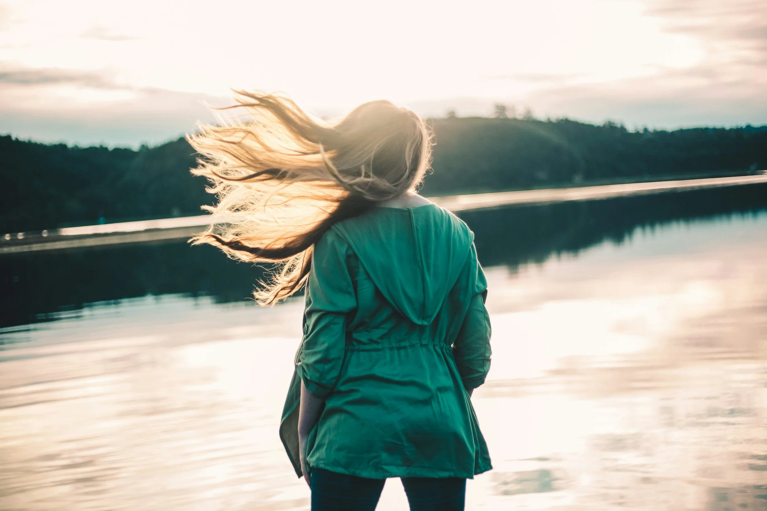 A woman with long hair wearing a green jacket standing near a body of water at sunset or sunrise.
