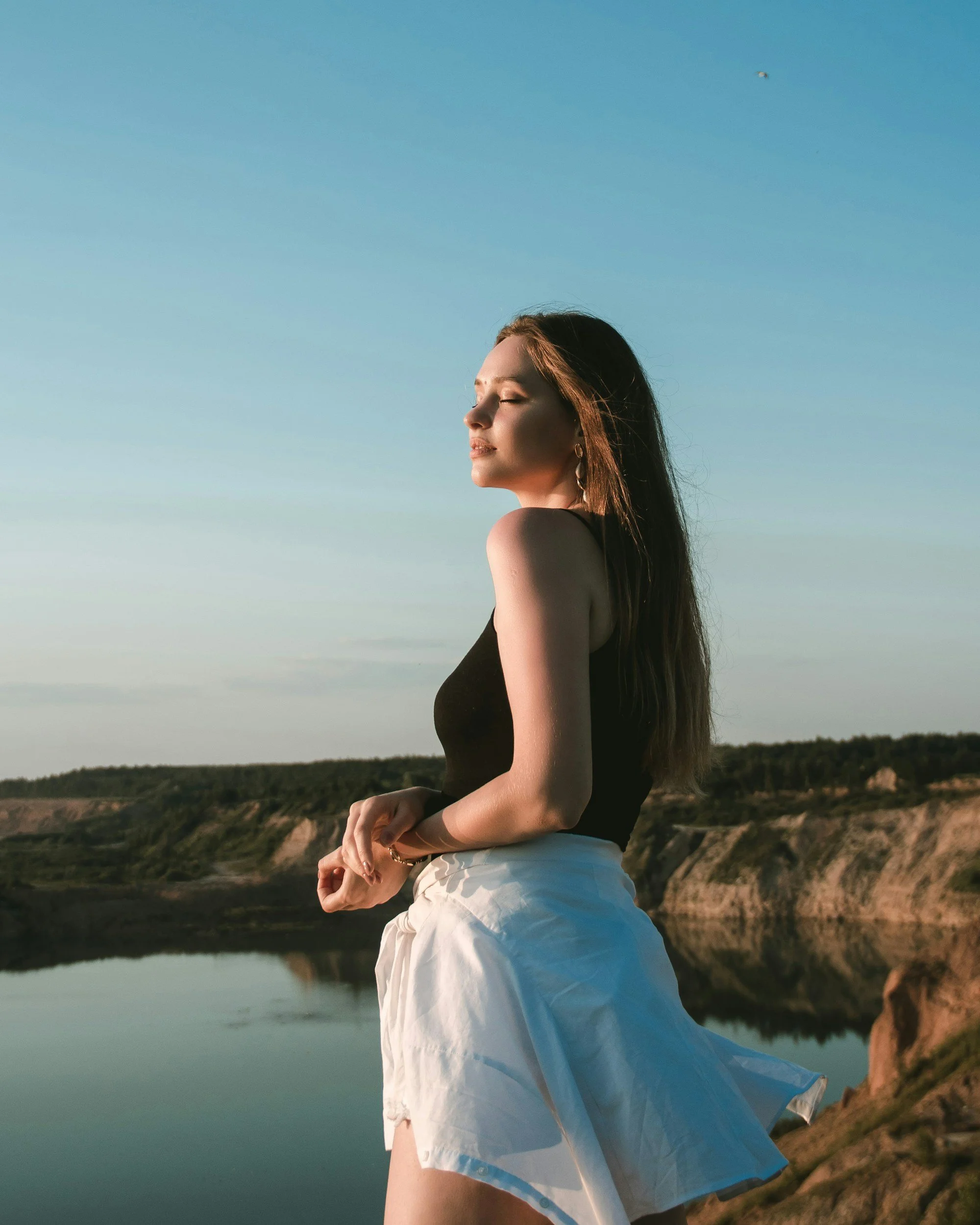 A woman stands outdoors with eyes closed, wearing a black top and a white skirt, in a natural landscape with water and hills in the background, during sunset or late afternoon.