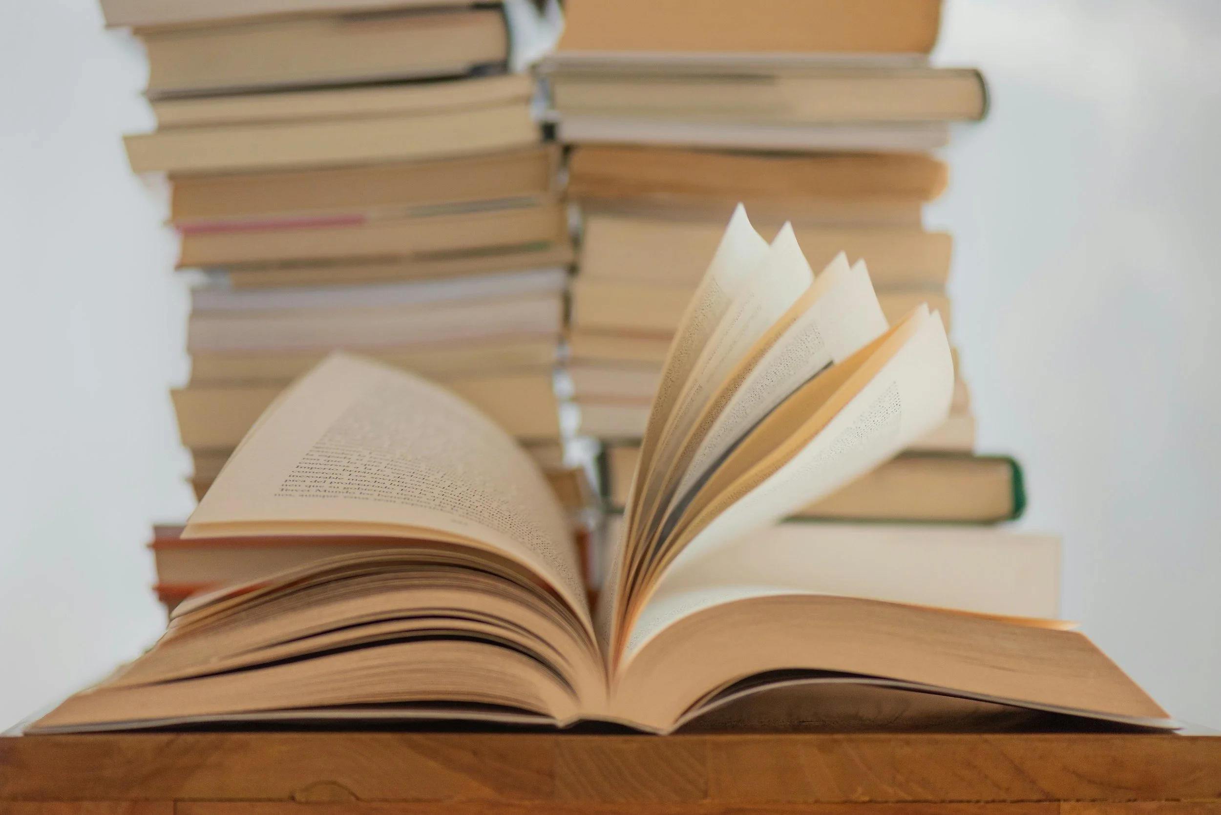 Open book with pages flipped on a wooden surface, with a large stack of closed books in the background.