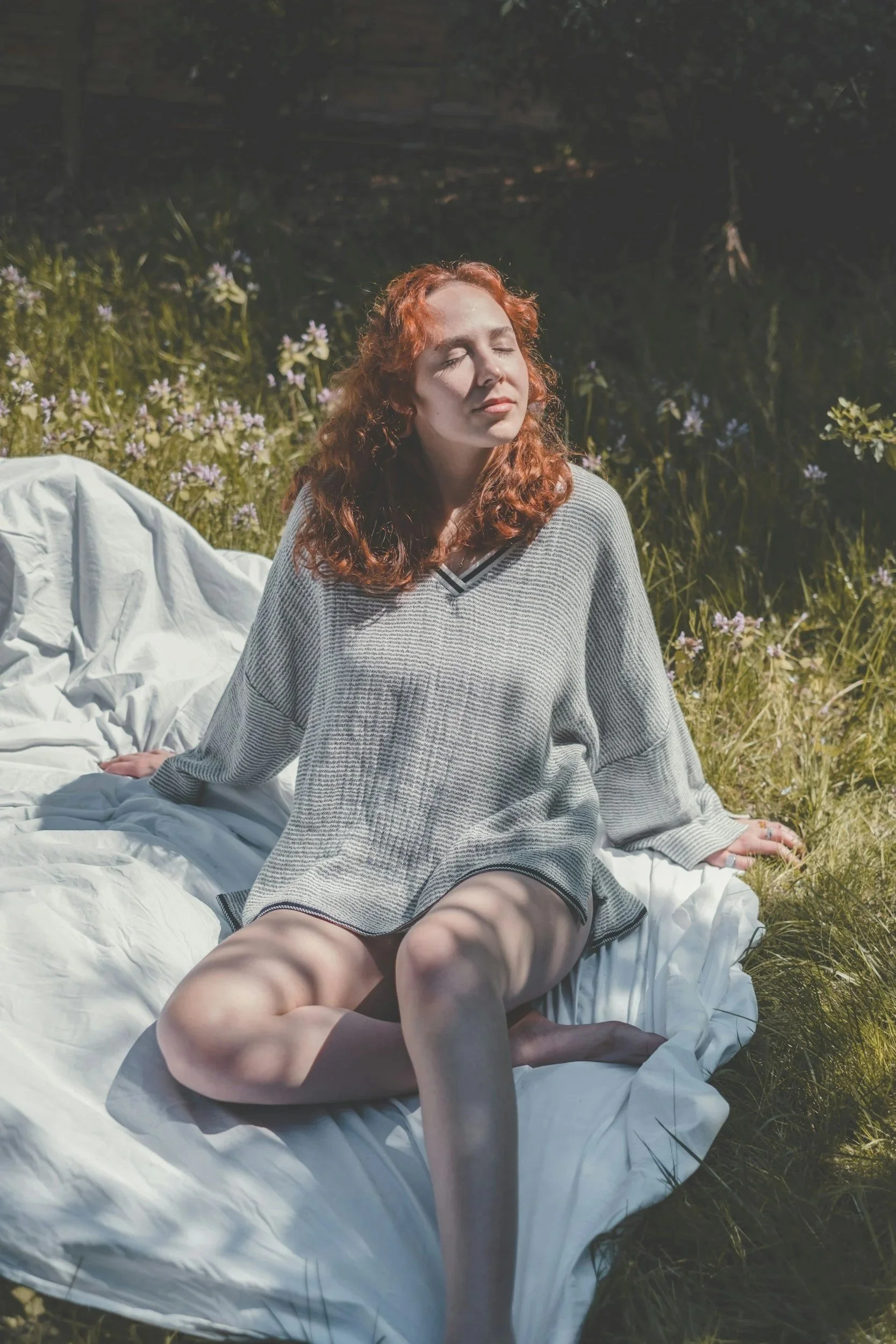 A woman with red curly hair and fair skin sitting outdoors on a white blanket among green grass and purple flowers, with her eyes closed, enjoying sunlight.