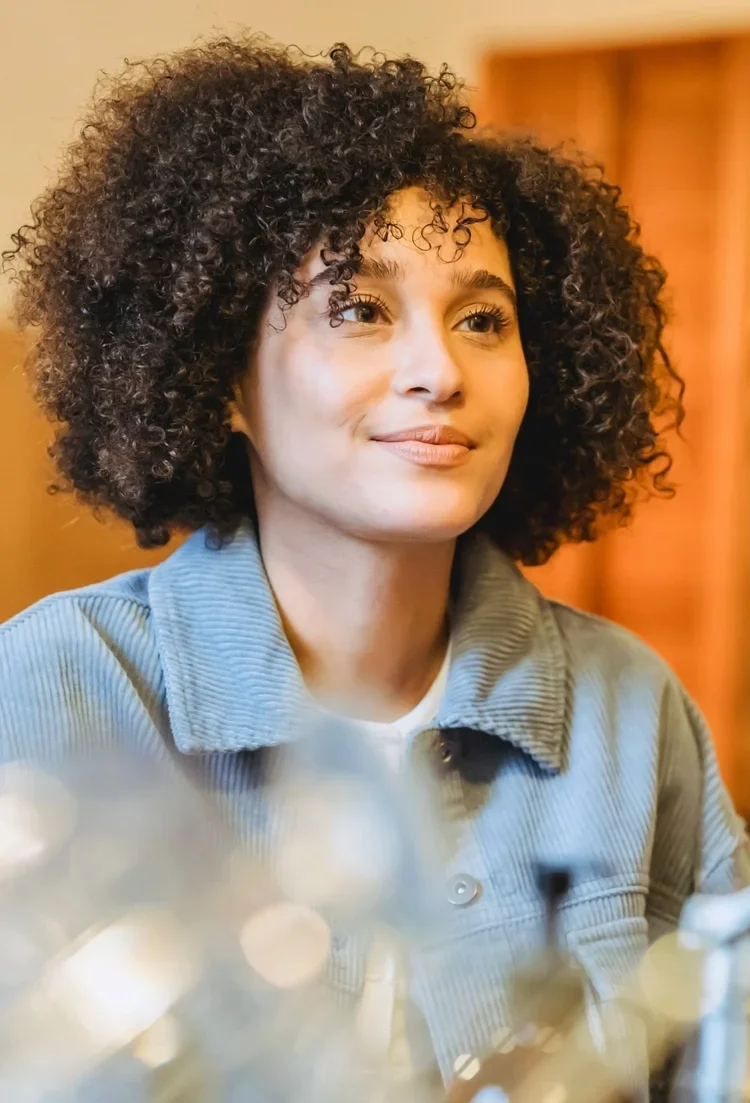 A woman with curly dark hair, wearing a light blue denim jacket, looking to her right with a slight smile, indoors with warm lighting.