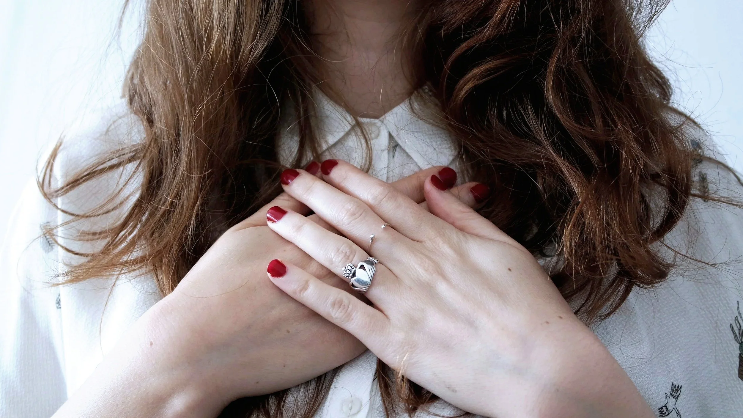 Woman with long wavy red hair, wearing a white shirt, holding her hands over her chest, showing rings and red nail polish.