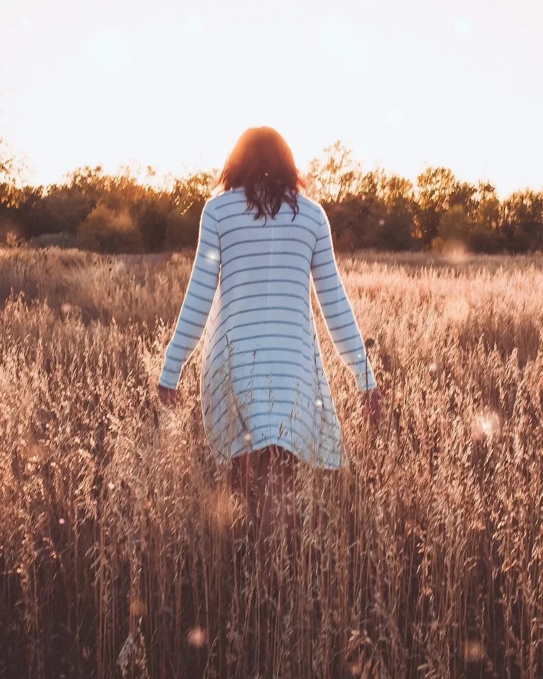 A woman walking through a field of tall grass during sunset, wearing a white dress with blue stripes.