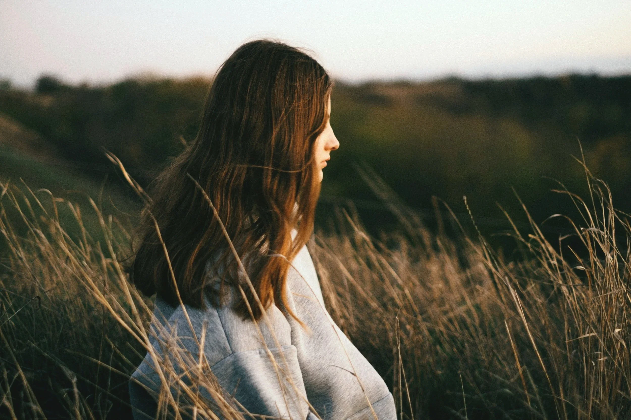 A woman with long brown hair standing in a field of tall golden grass, facing sideways during sunset or late afternoon.
