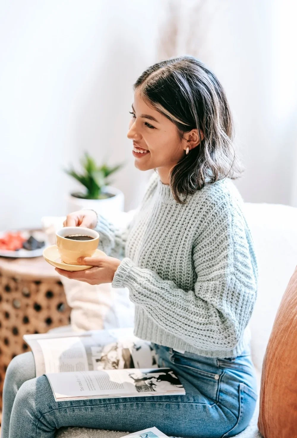 A young woman with shoulder-length dark hair, smiling, sitting on a beige chair, holding a yellow coffee cup and saucer, wearing a gray knitted sweater and blue jeans, with an open magazine on her thighs, in a bright, cozy room with a plant and a table with snacks in the background.