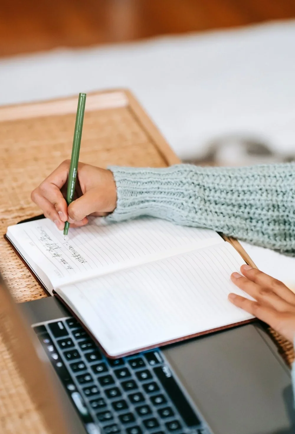 Person writing in a notebook with a green pen, seated at a wooden desk with a laptop nearby.