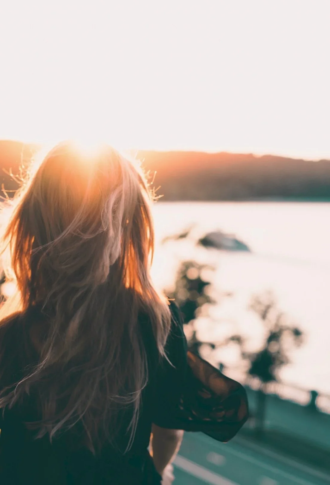 A woman with long hair standing outdoors near a body of water during sunset.