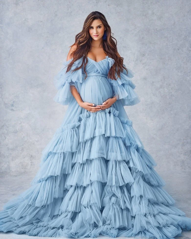 A pregnant woman in a flowing, tiered, light blue gown with ruffles, standing against a neutral background, holding her belly with both hands.