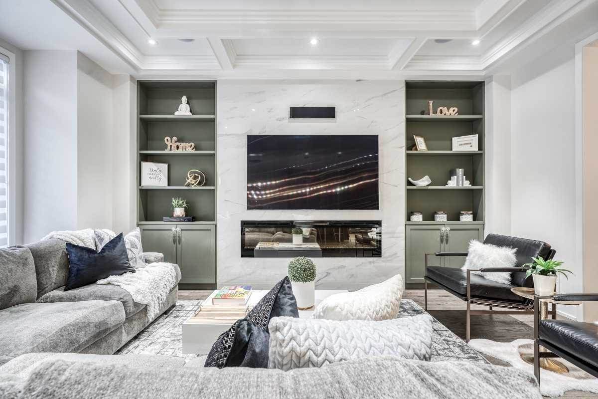 Modern living room with gray and black furniture, a large flat-screen TV mounted on a white marble wall, and built-in shelves with decorative items and books.