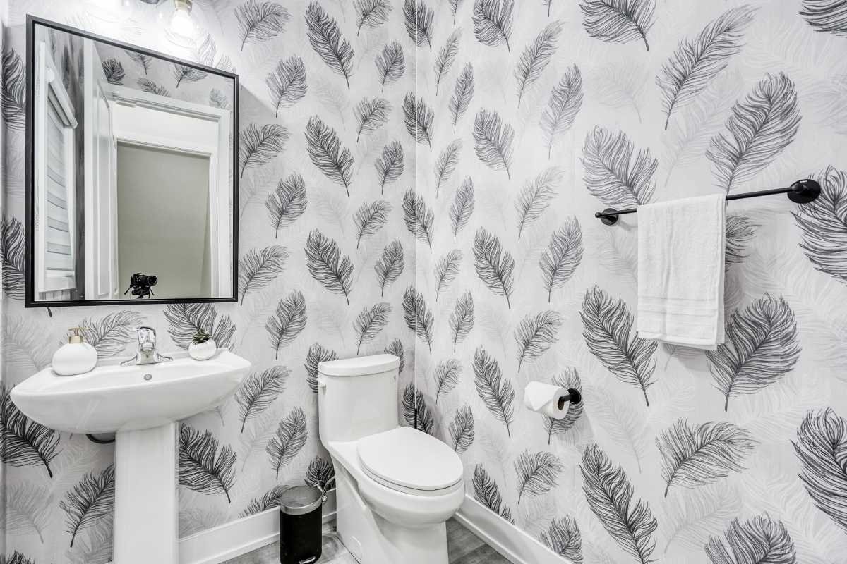 Bathroom with wallpaper featuring black and gray feathers, a white pedestal sink with decorative items, a wall-mounted mirror, a white toilet, a black trash can, a towel on a black towel bar, and a toilet paper roll.