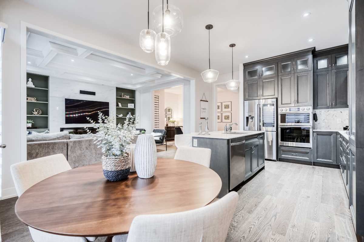 Open concept kitchen and living room with modern decor, gray cabinets, stainless steel appliances, a wooden dining table with vases, and a cozy seating area with a wall-mounted TV.