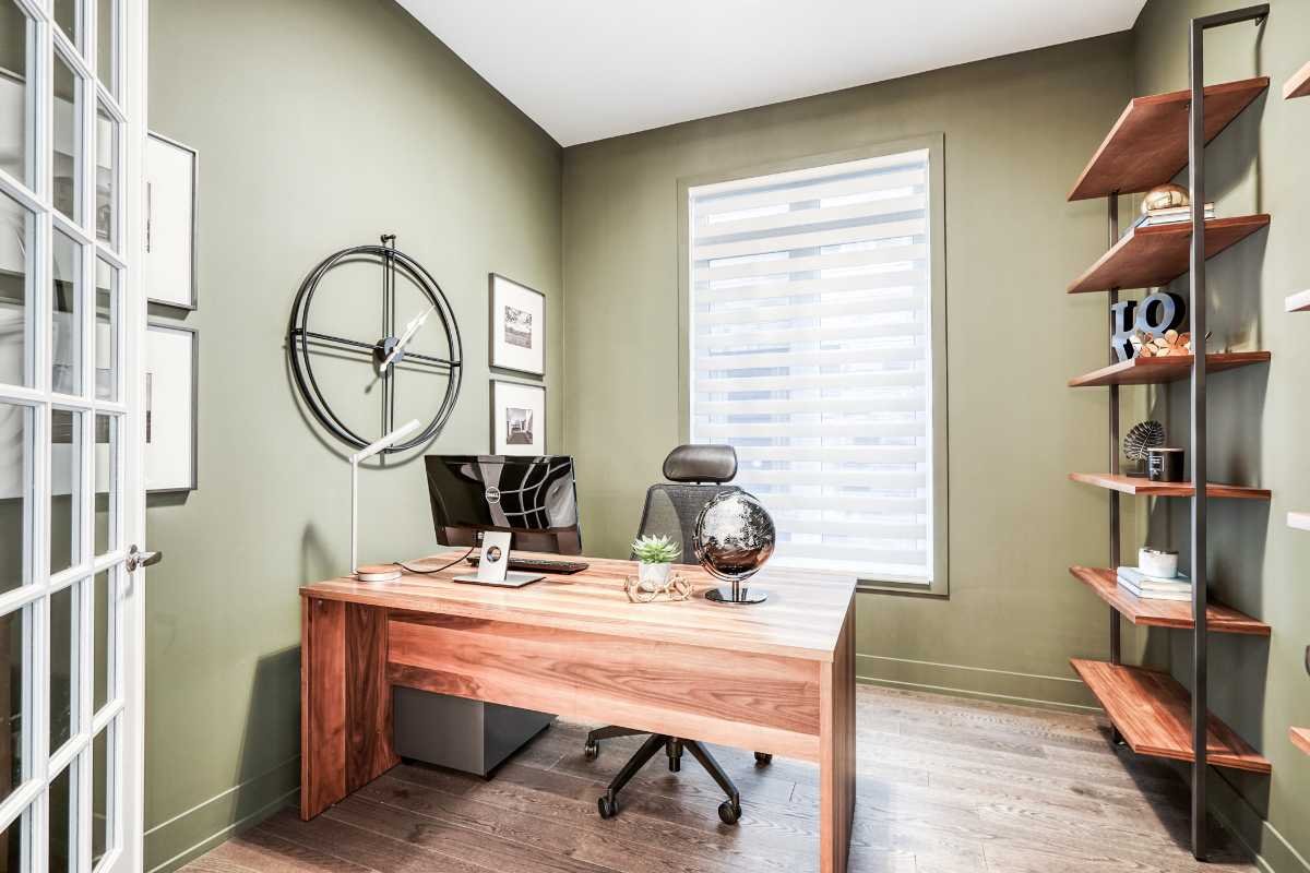 A modern home office with green walls, a wooden desk with a computer, decorative items, a black office chair, and a shelf with decor, books, and a clock.