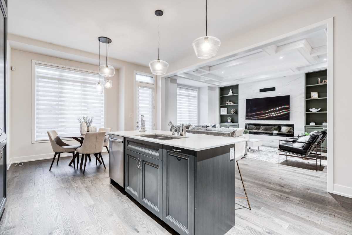 Modern open-plan living and dining area with a kitchen island, dining table with six chairs, and a living room with a wall-mounted TV, built-in shelves, a white sectional sofa, and black accent chairs. Bright natural light streams through large windo