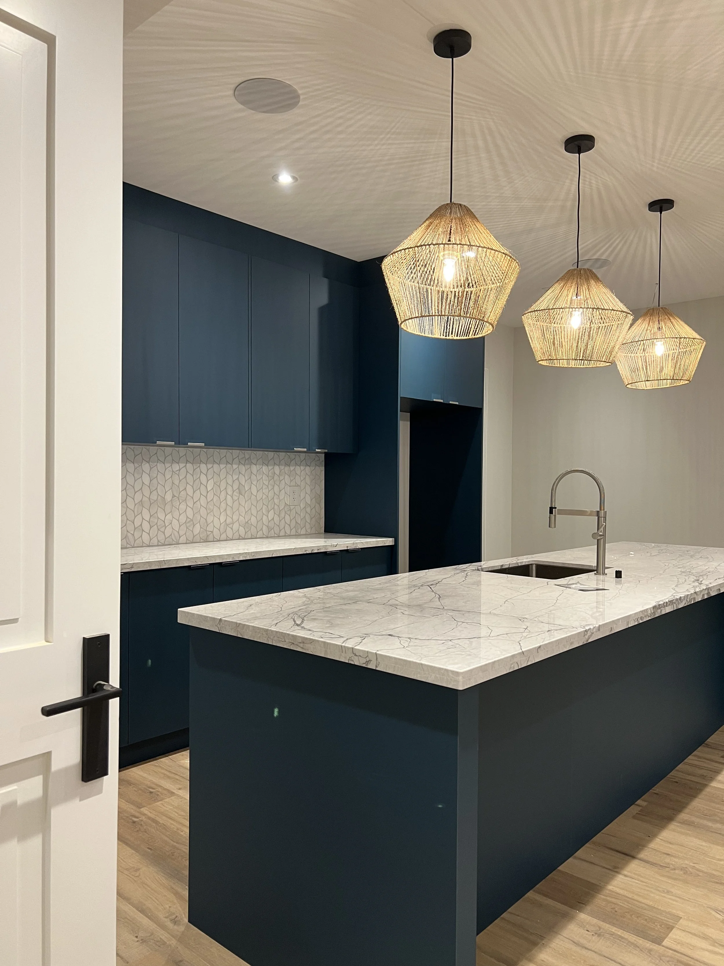 Modern kitchen with white marble island, blue cabinets, geometric backsplash, and three woven pendant lights.