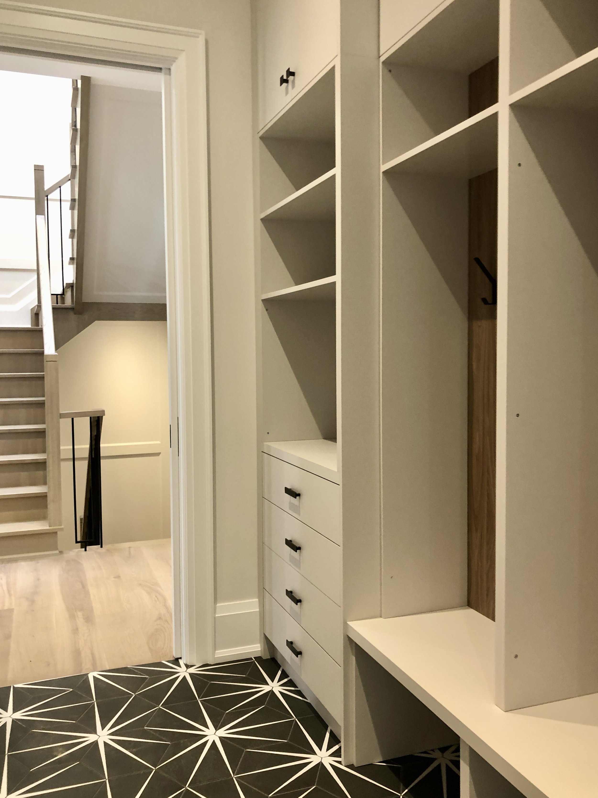A hallway with built-in white shelving and drawers, black hardware, and a geometric black and white tile floor near a staircase with wood steps and a black metal railing.