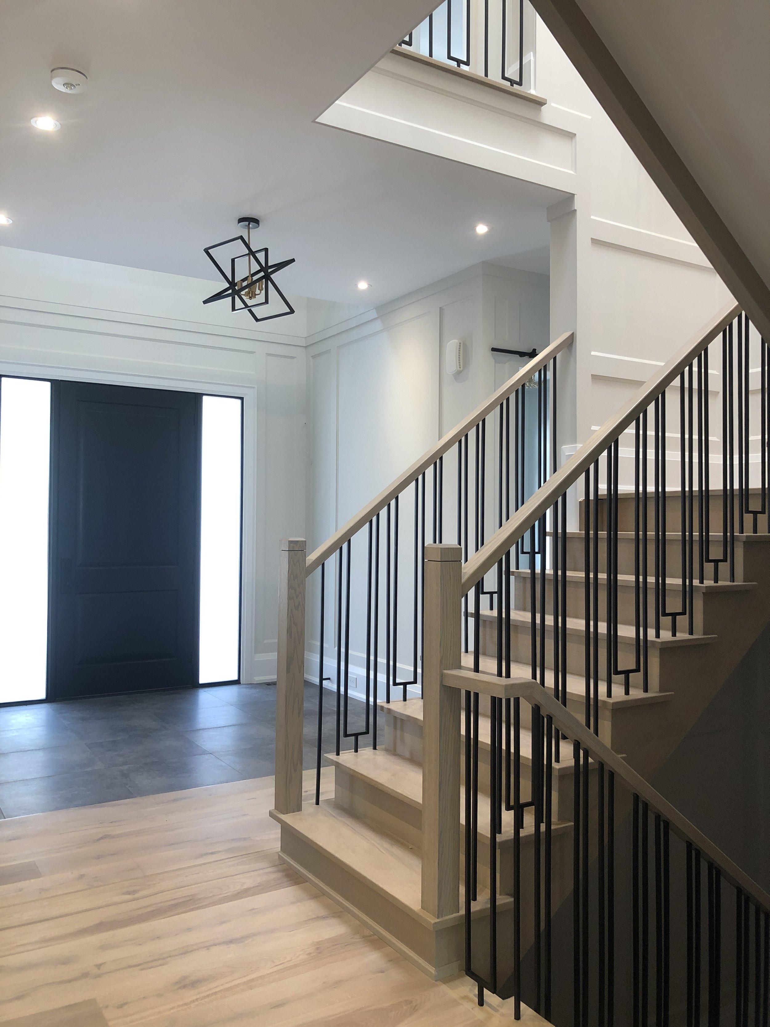 Interior view of a modern home staircase and entryway, with a wooden staircase featuring black metal railings leading upstairs, a black front door with glass panels, and a ceiling light fixture with geometric design.
