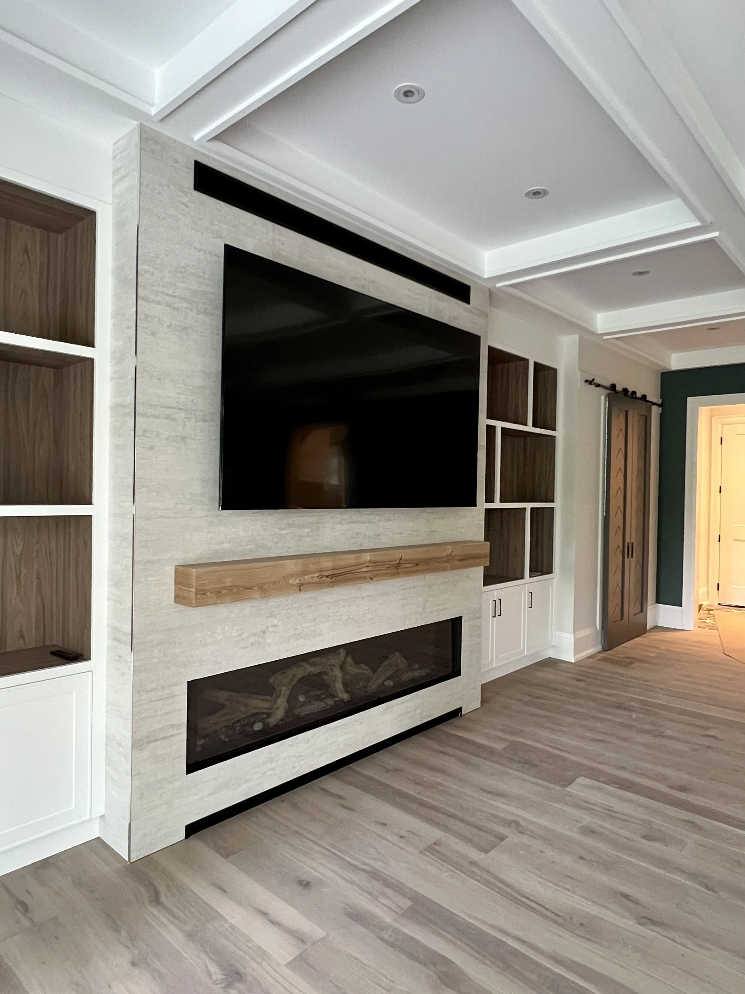 Modern living room with wall-mounted flat-screen TV, electric fireplace, and built-in shelves, with wooden flooring and a tray ceiling.