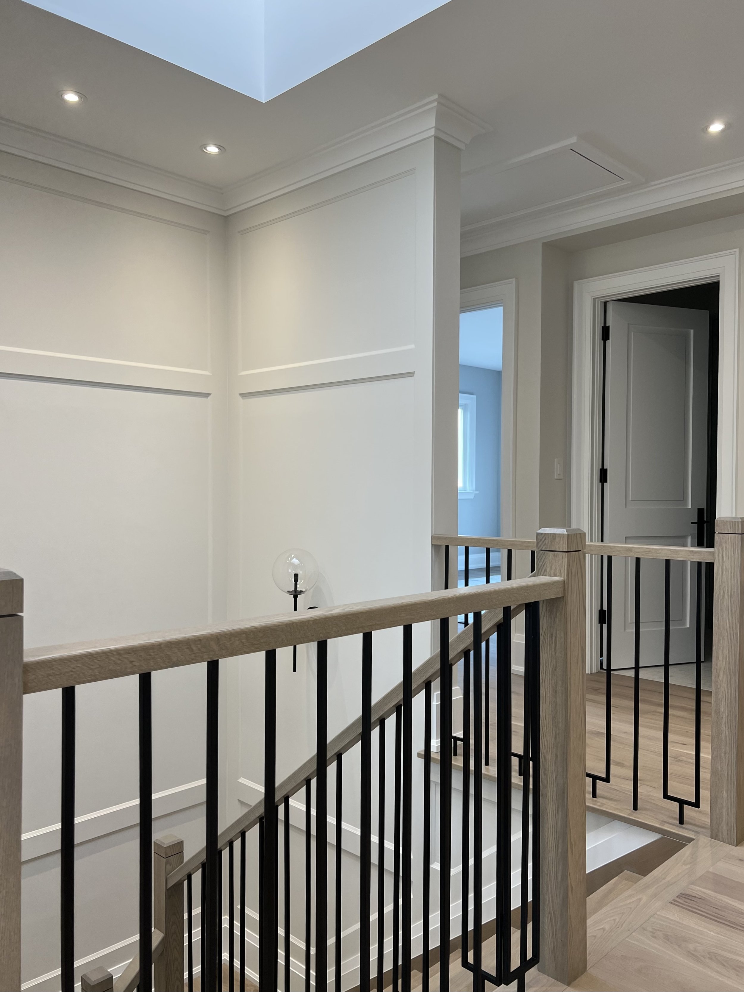 Indoor staircase with wooden handrail and black metal balusters leading down to the lower level in a modern home, with white walls and ceiling, recessed lighting, and two open doors visible in the background.